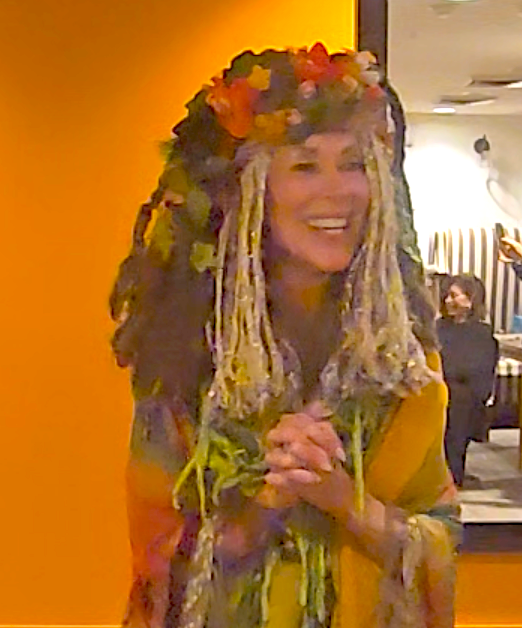 Woman in floral headdress and shawl, smiling, hands clasped, near an orange wall.
