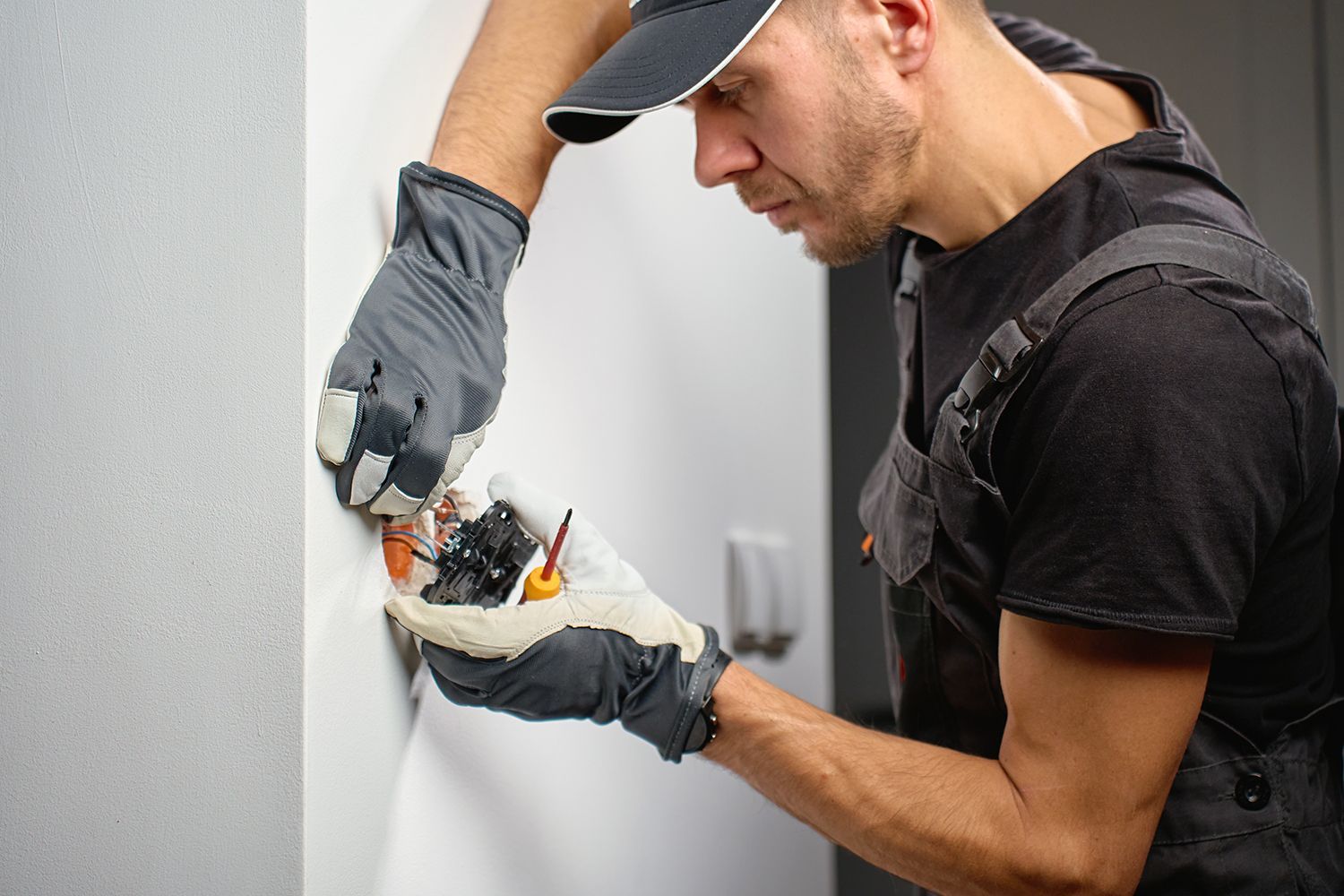 Worker in gray gloves installing electrical wiring inside a wall, holding tools and cables.