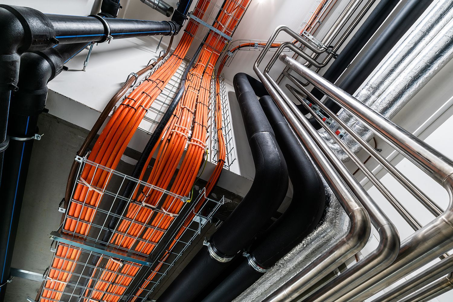 Overhead view of orange and silver electrical cables in metal cable trays and conduits running through a ceiling space