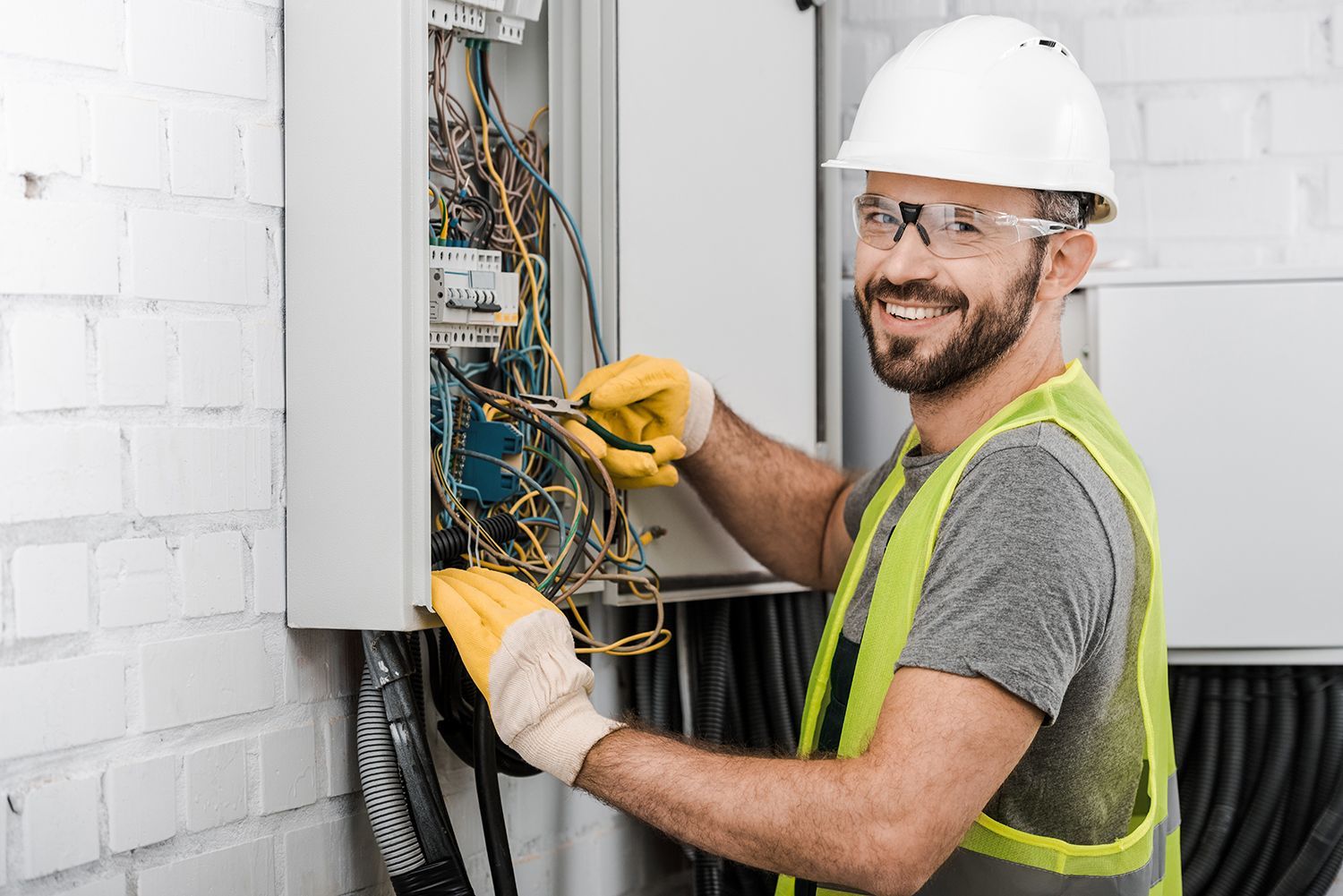 Electrician in hard hat wiring an open electrical panel, wearing gloves and a safety vest. Electrician in hard hat wiring an open electrical panel, wearing gloves and a safety vest.