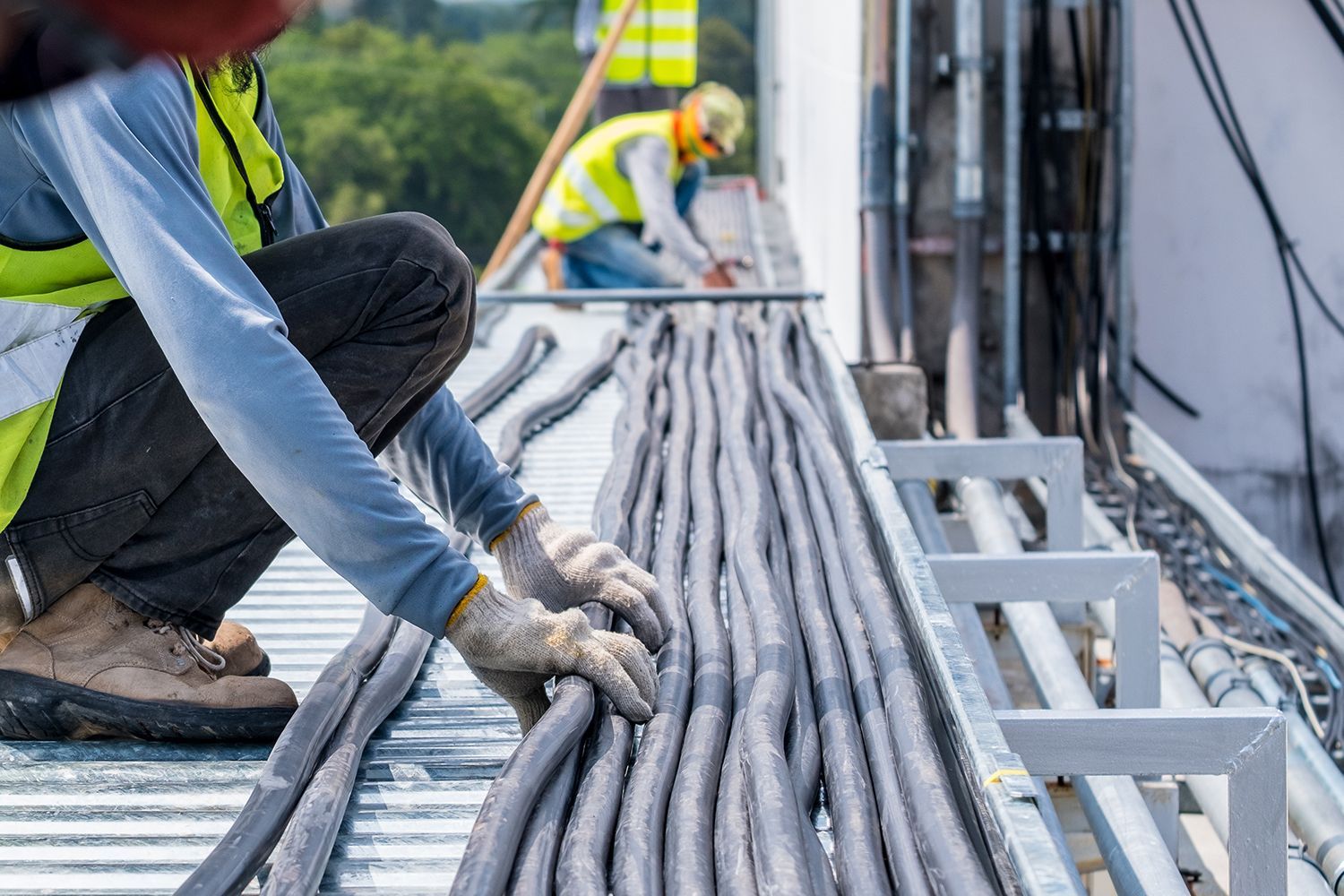 Workers handling thick cables on a construction site, with a person kneeling in gloves and a safety vest.