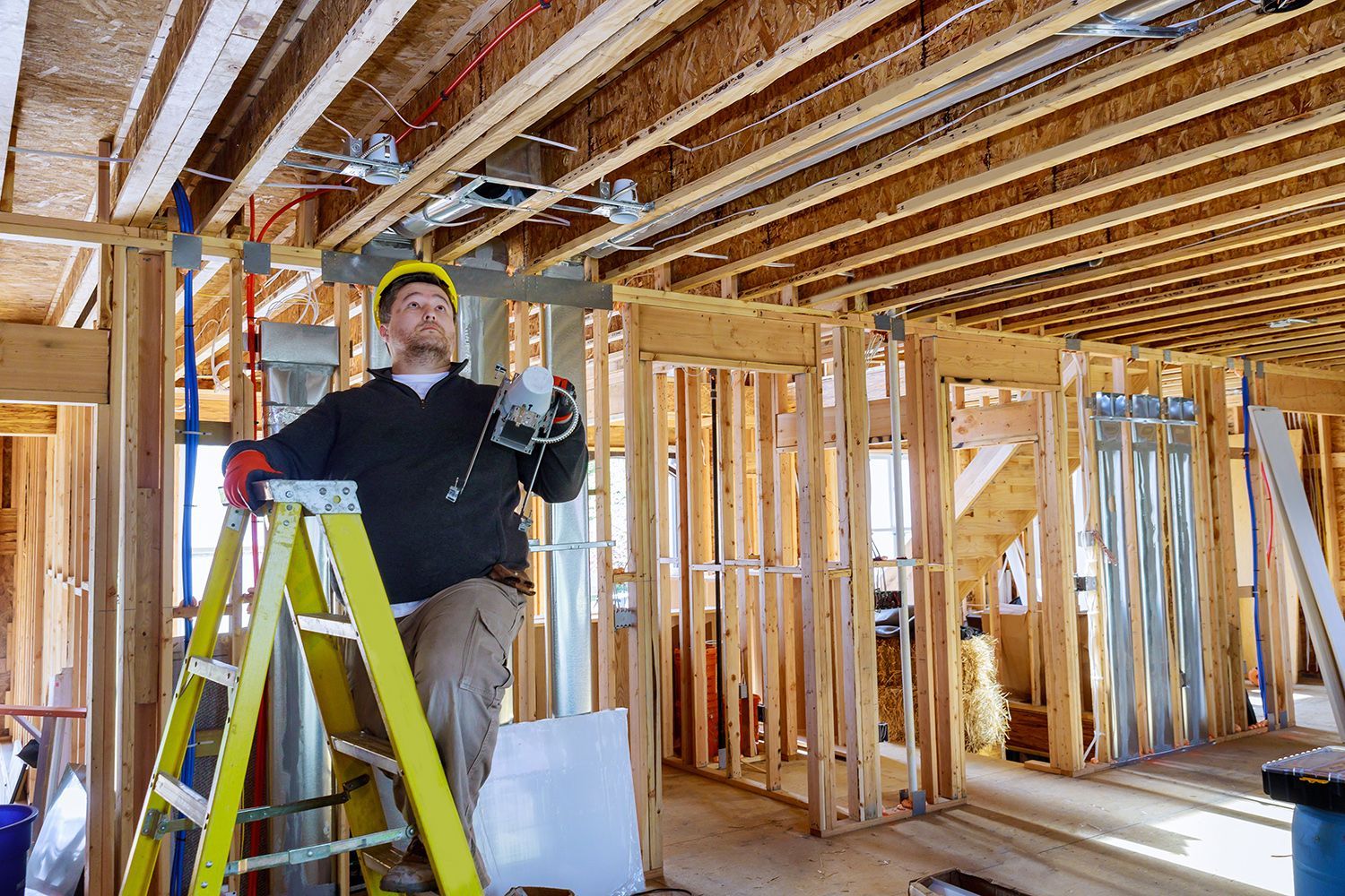 Person standing on a yellow ladder inside a framed house under construction.