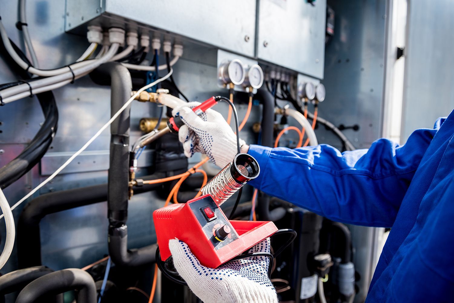 Technician in blue workwear using a red meter to test electrical equipment inside a control panel