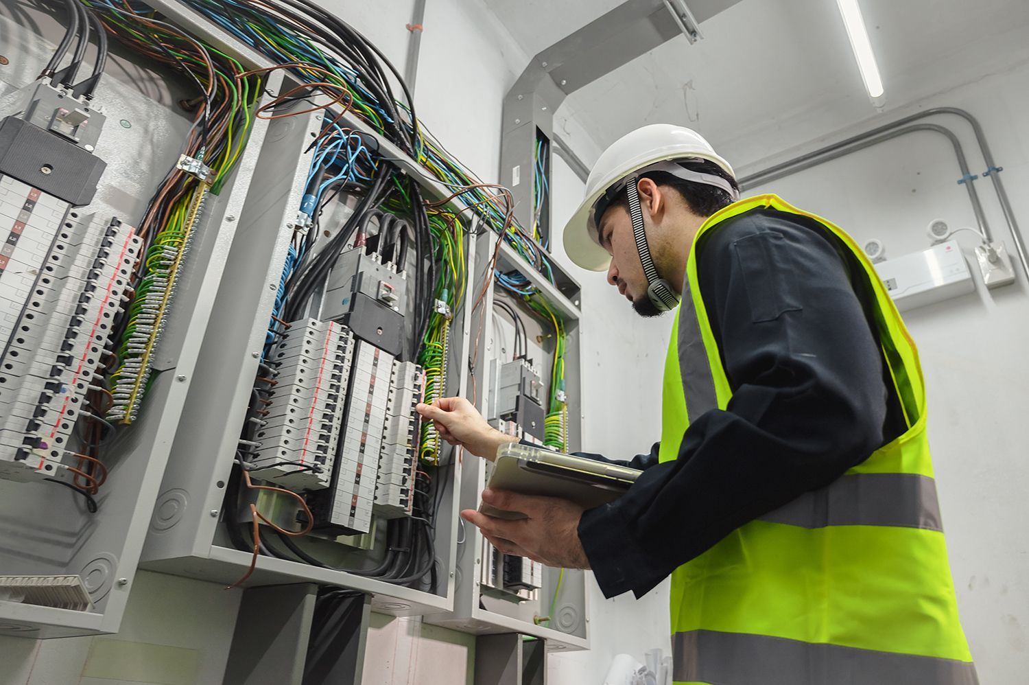 Technician in a hard hat and safety vest inspecting wiring inside an electrical control panel room Technician in a hard hat and safety vest inspecting wiring inside an electrical control panel room