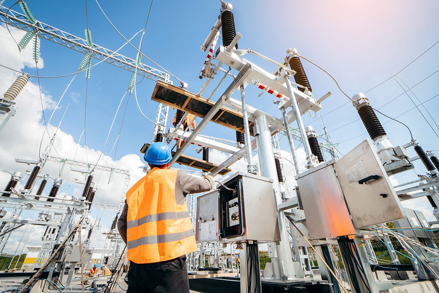 Worker in orange safety vest inspecting electrical substation equipment under a bright sky
