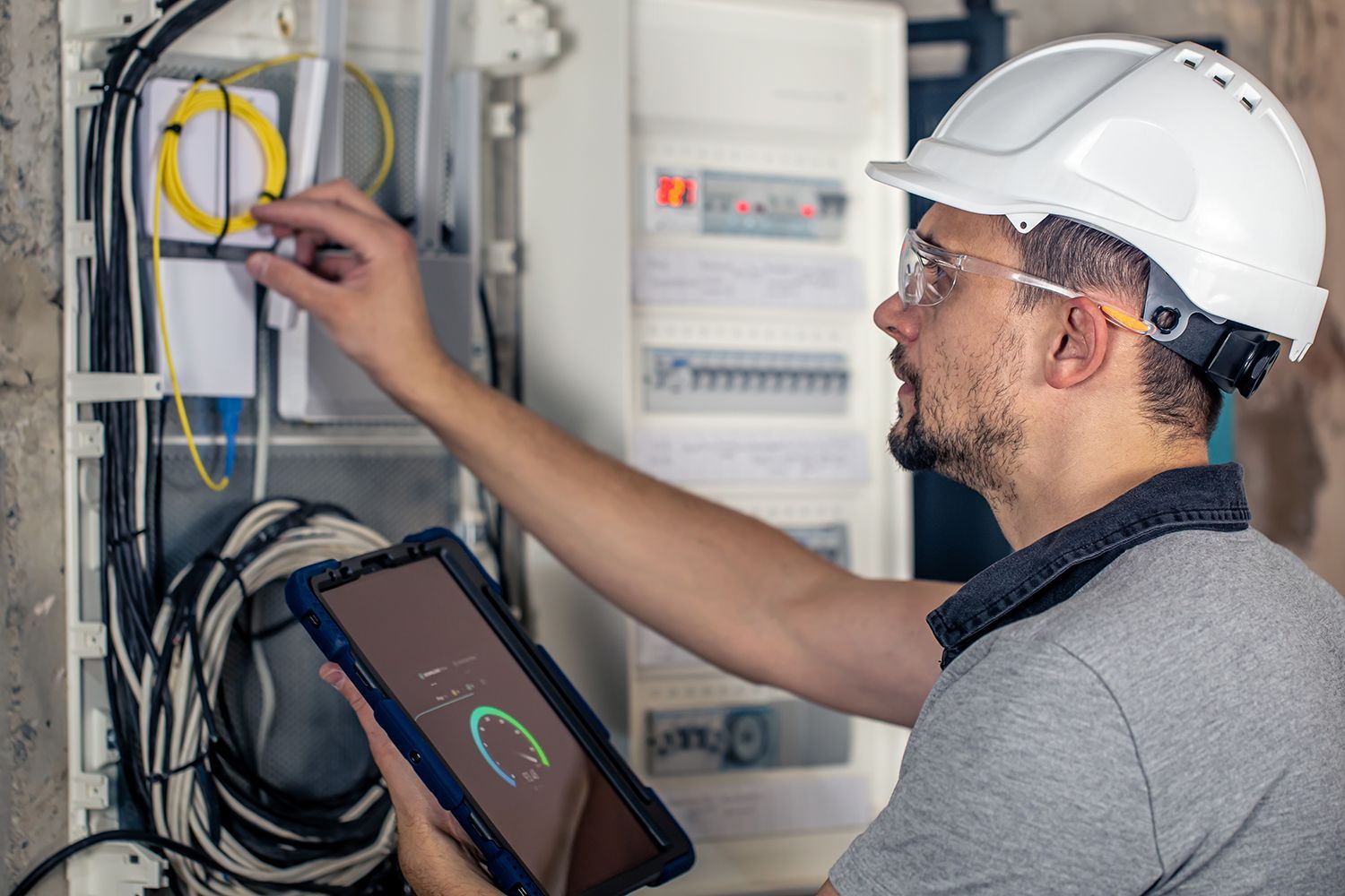 Technician inspecting electrical panel with tablet in a utility room, wearing hard hat and safety glasses