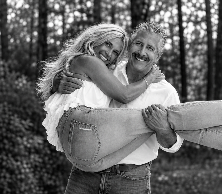 Smiling couple outdoors, woman hugging man from a burlap sack hammock in a wooded setting
