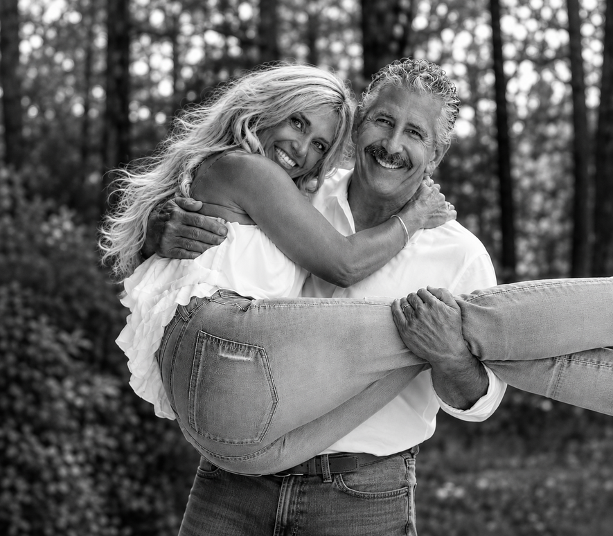 Smiling couple hugging outdoors, with the woman lifted in the man's arms against a tree-lined background.
