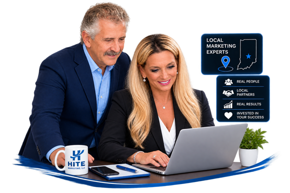 Two professionals reviewing a laptop at a desk, with a coffee mug and marketing graphics beside them