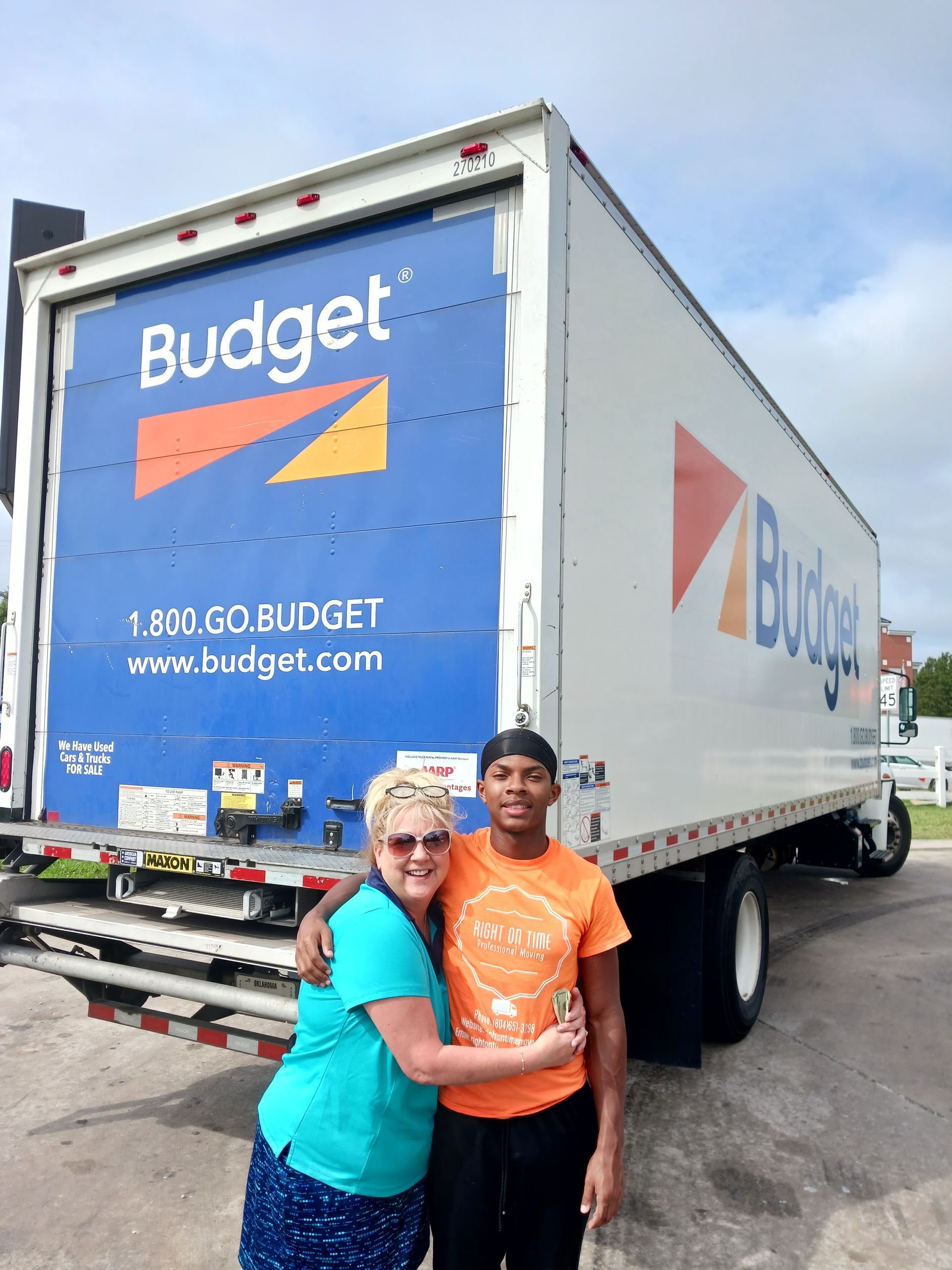 Two people posing beside a blue Budget moving truck in a parking lot.