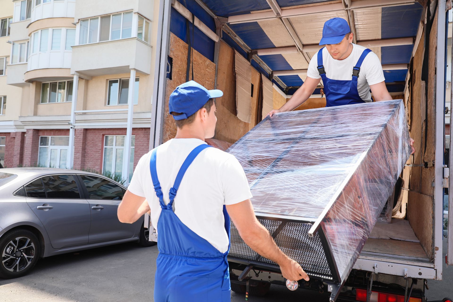 Two movers in blue overalls unloading a wrapped sofa from a truck on a city street