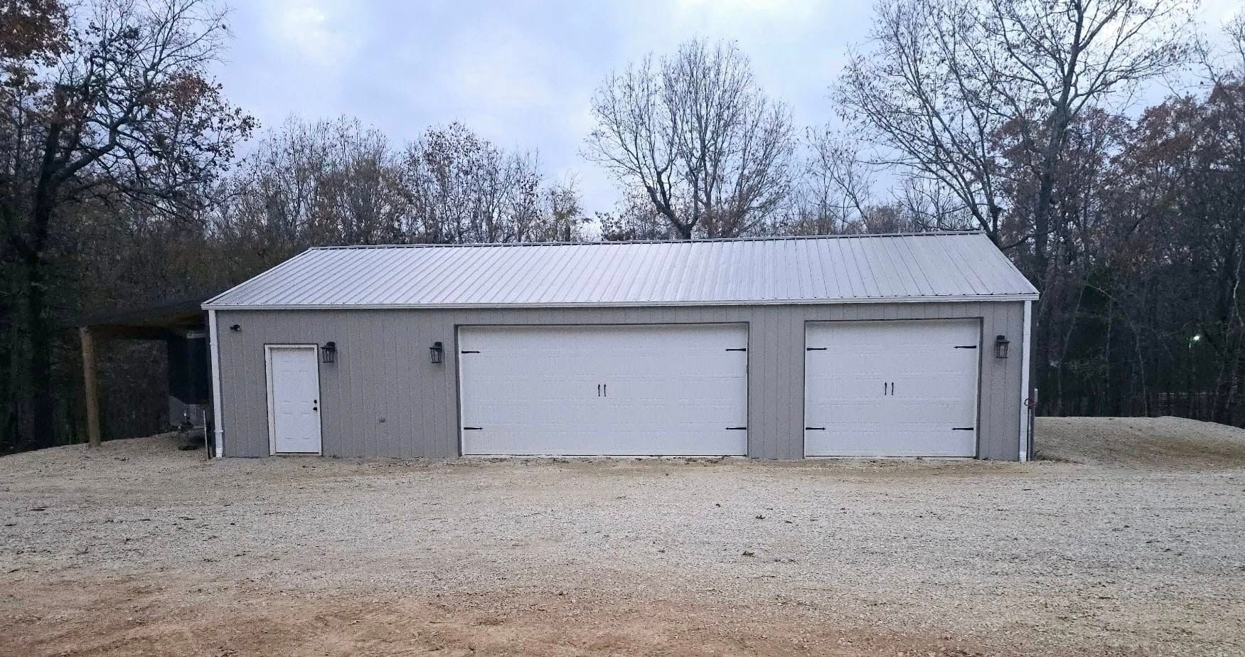 A gray metal garage with three doors sits on gravel with bare trees in the background.