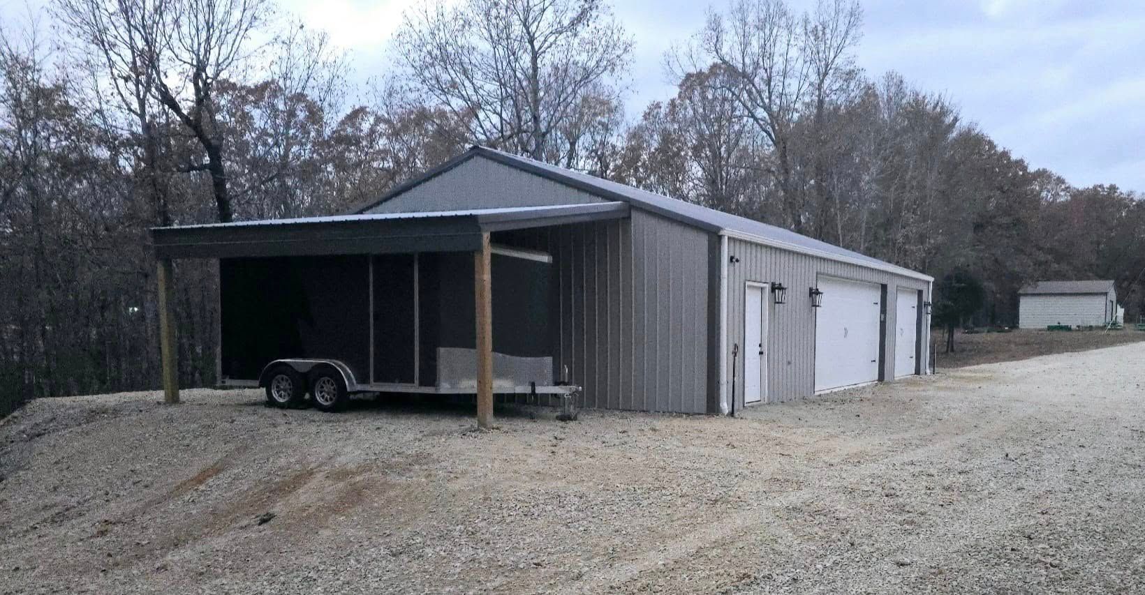 Metal building with a covered awning, gravel driveway, trailer parked under awning.
