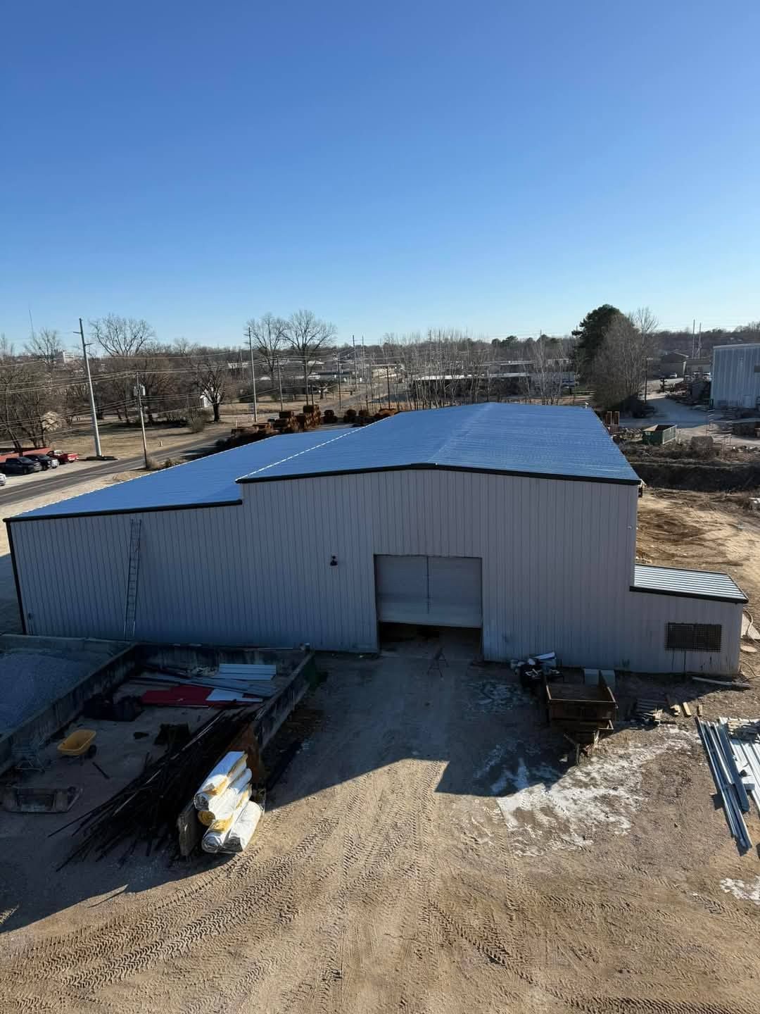 Large industrial building with blue roof under a clear, sunny sky. Construction materials and debris in the foreground.