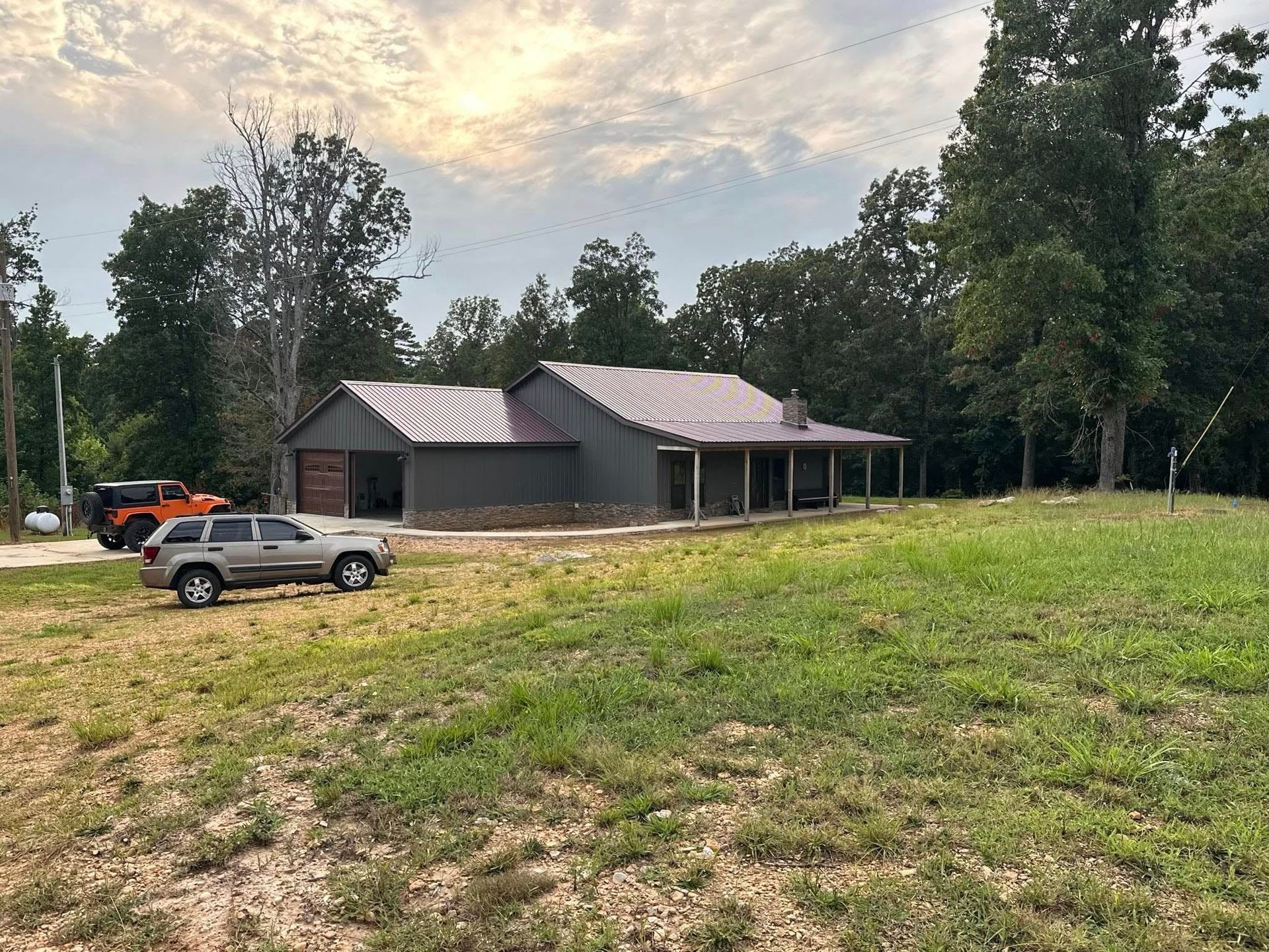 A house with a dark metal roof, cars parked outside, and a grassy yard under a cloudy sky.