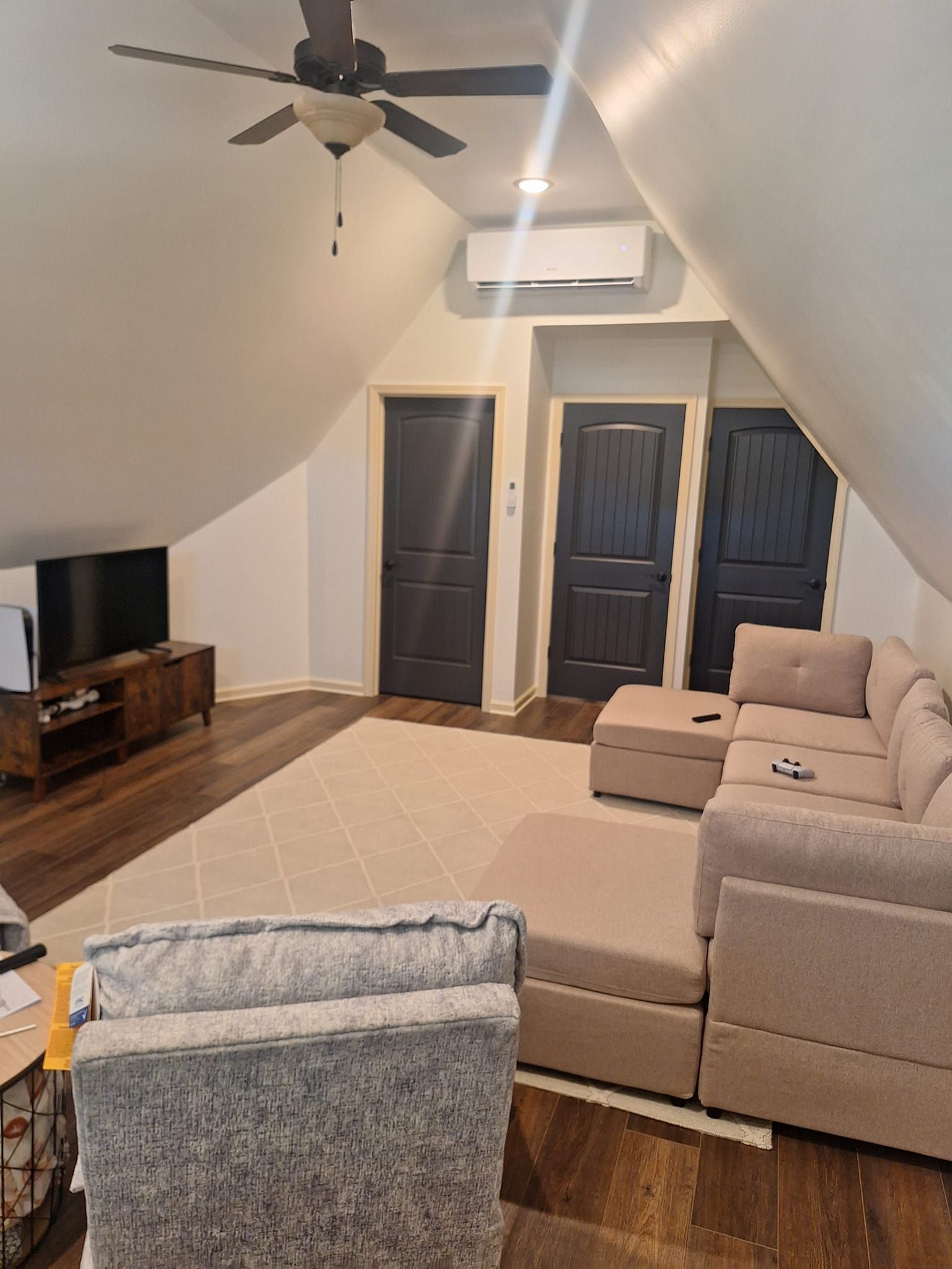 Living room with sloped ceiling, TV, sofa, and dark doors. Beige and brown tones throughout.