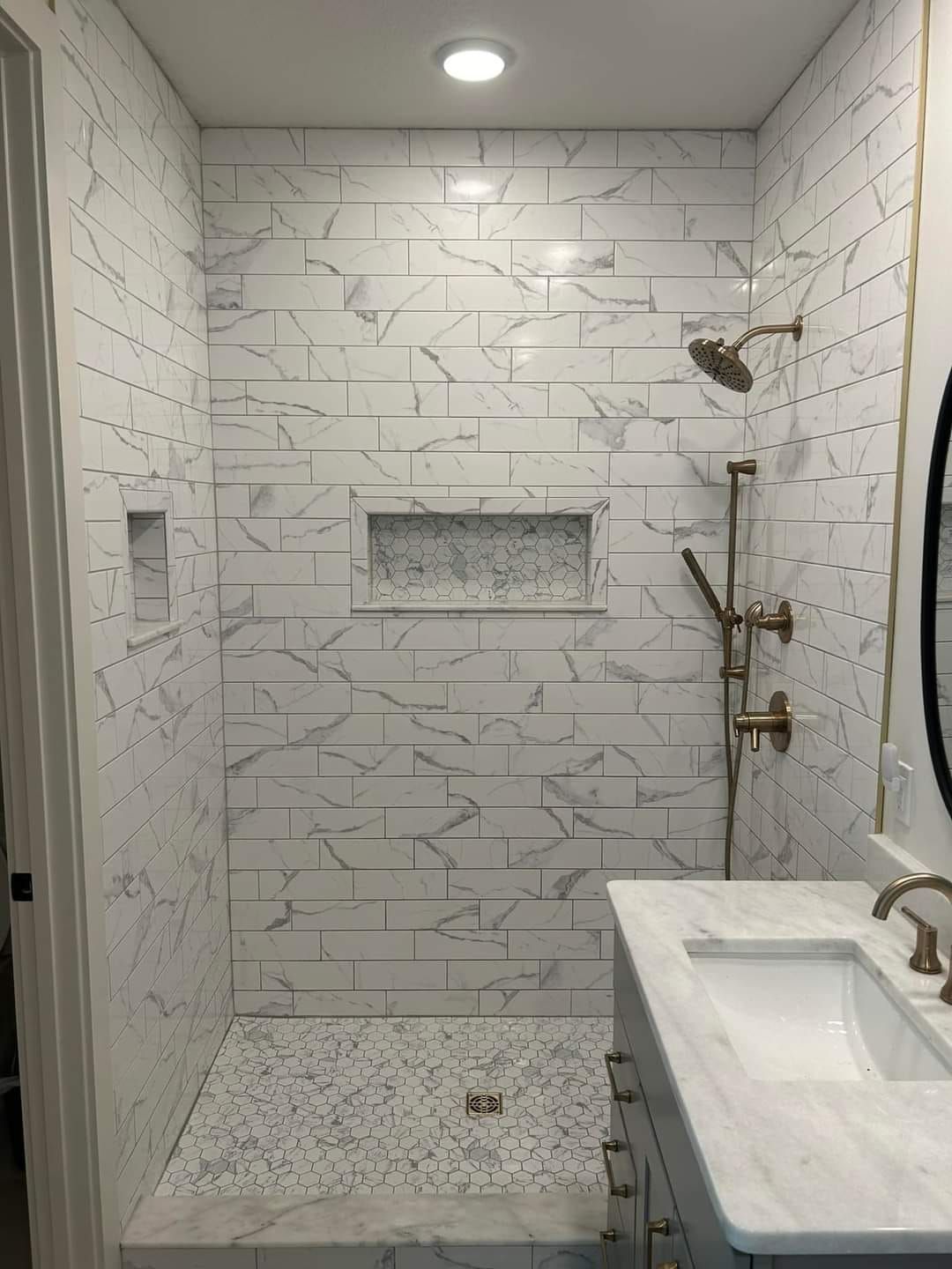 Modern bathroom with marble-look tiled shower and sink. Features include a built-in shelf, showerhead, and gold fixtures.