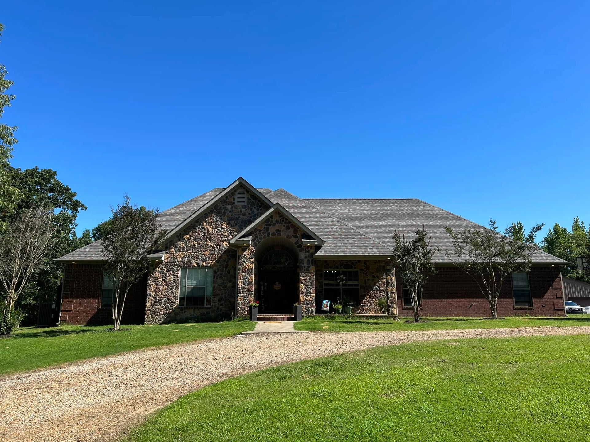 A single-story brick house with a stone facade, gray roof, and gravel driveway under a blue sky.