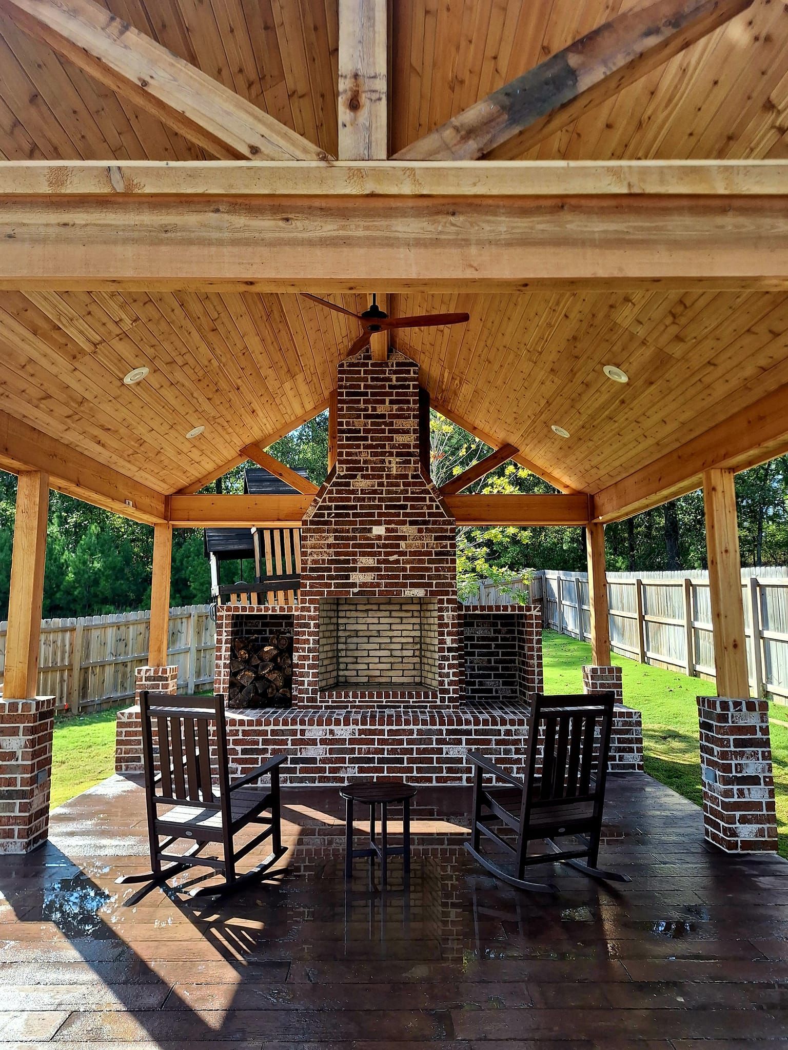 A wooden gazebo with a brick fireplace and rocking chairs