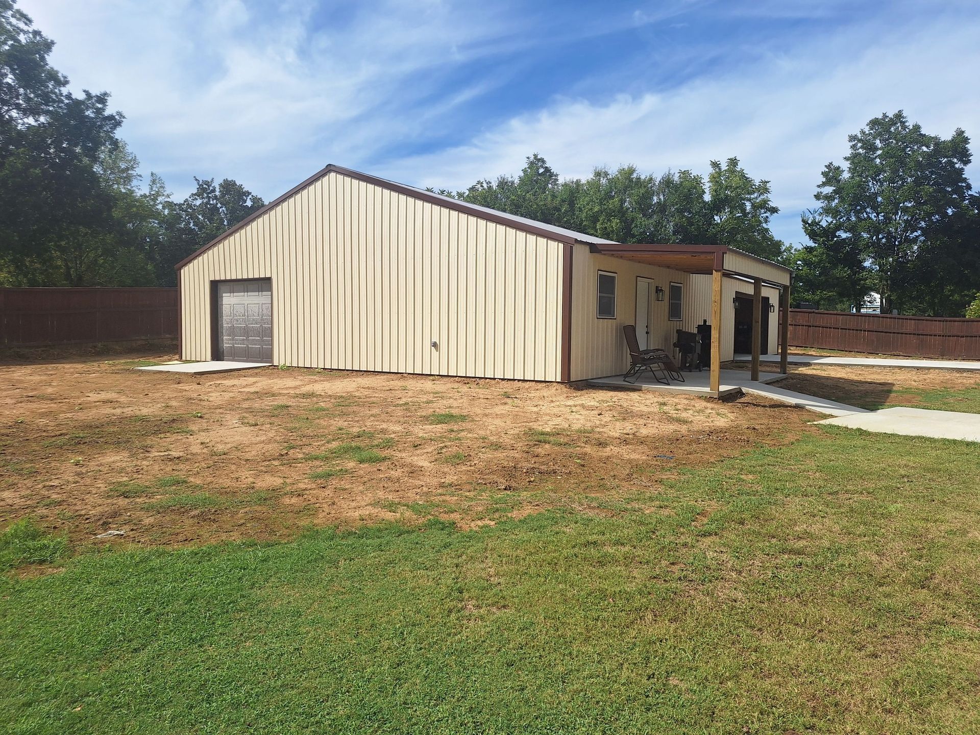 Tan metal-sided building with garage door, patio, and brown trim on a grassy lot under blue sky.