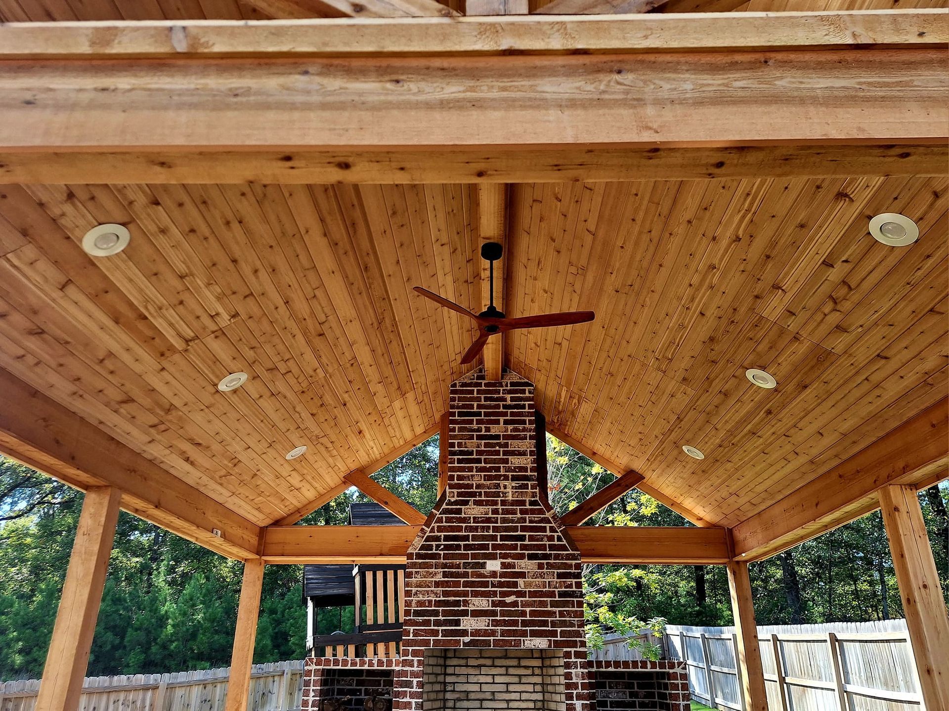 Wooden outdoor patio ceiling with brick fireplace and ceiling fan.