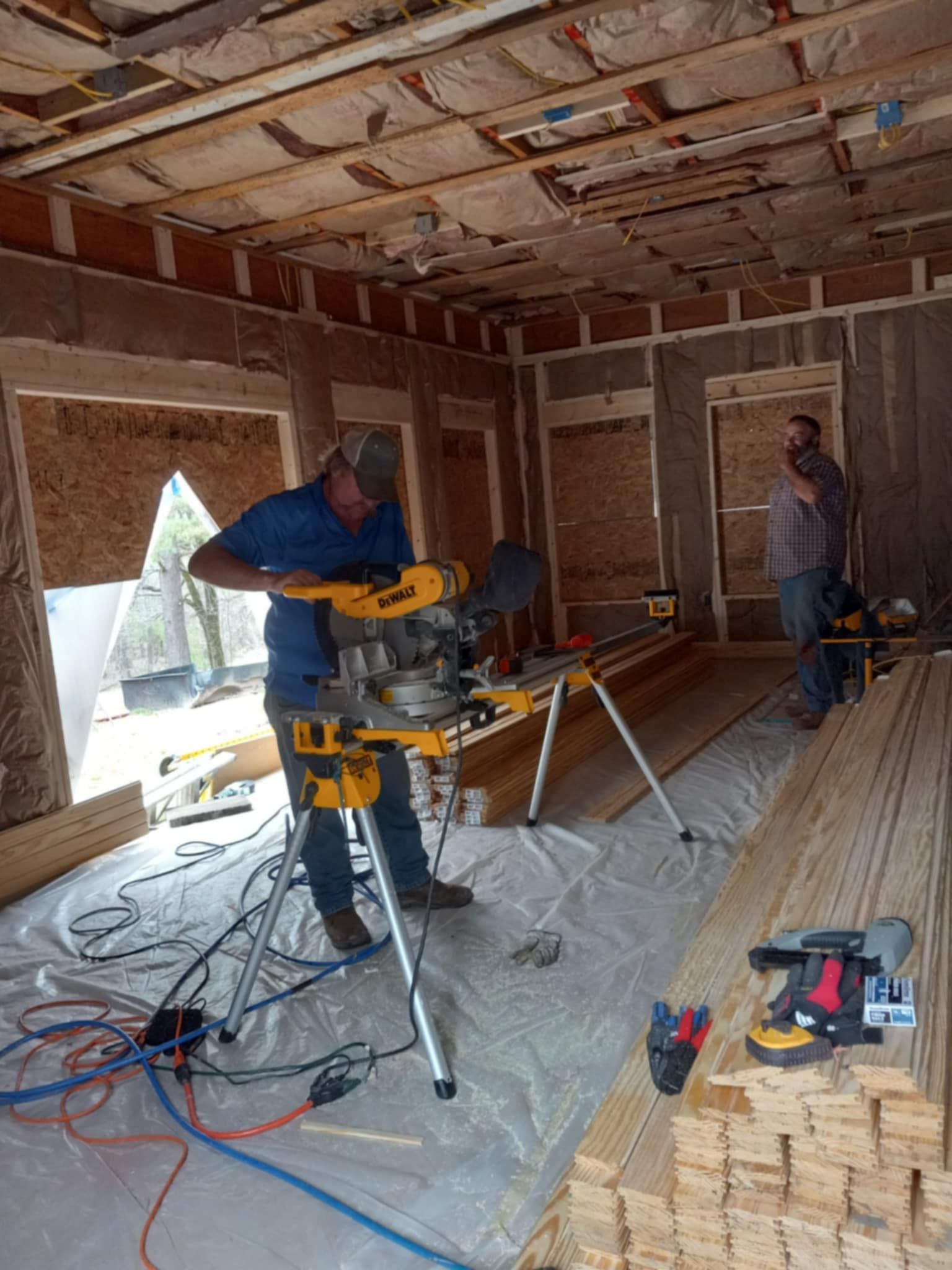 Two men working on a construction site. One uses a miter saw, the other observes. Interior framing is visible.