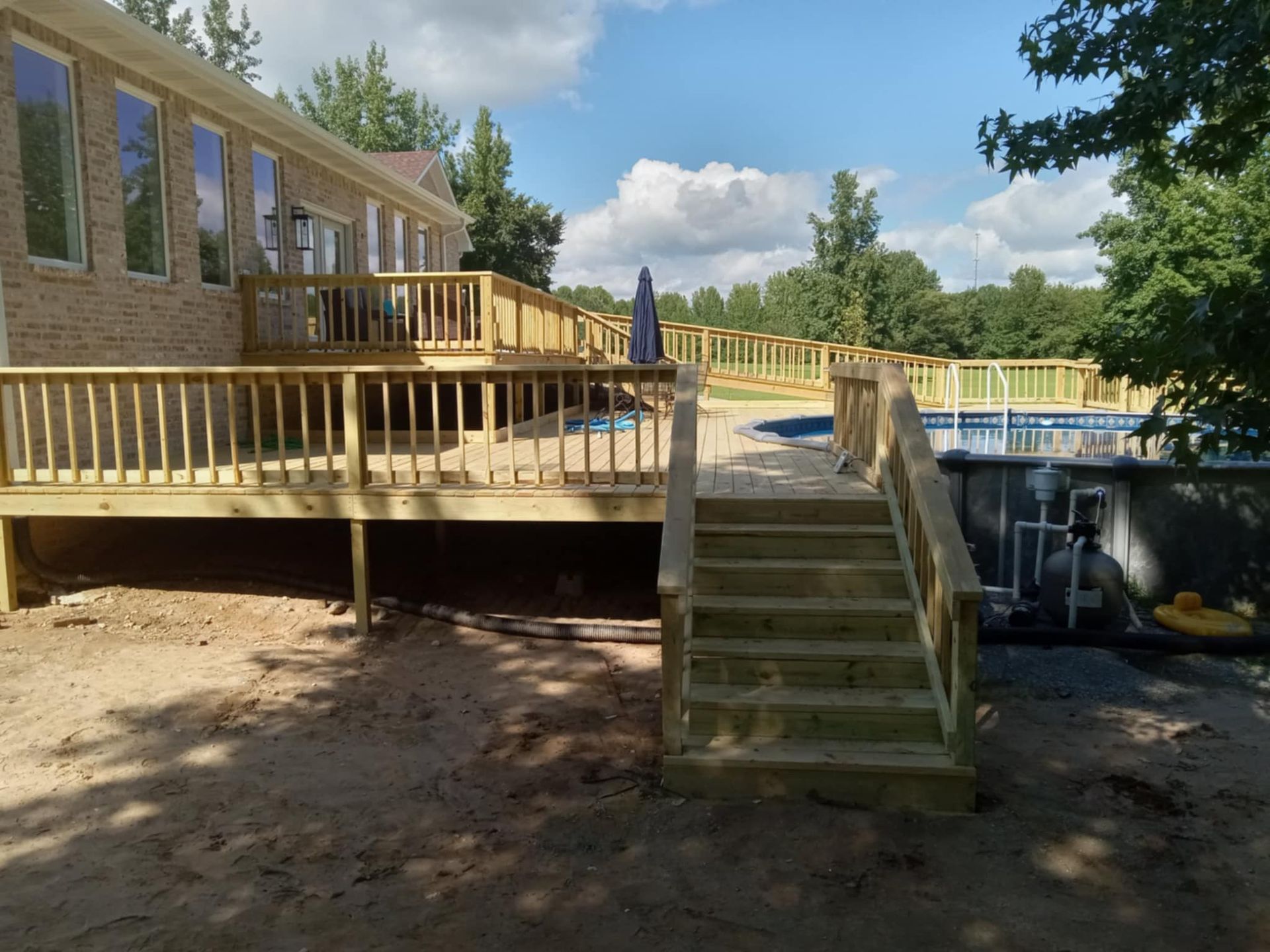 Wooden deck with stairs leads to above-ground pool; house with brick facade in the background. Sunny day.