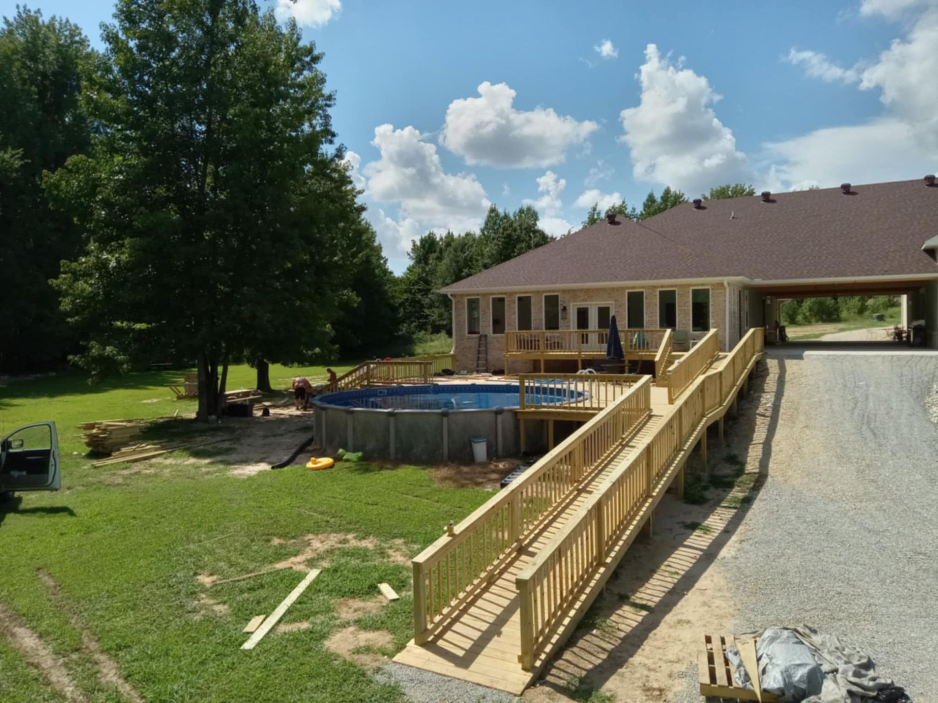A wooden ramp leads to a deck with an above-ground pool. A house with a brown roof is in the background on a sunny day.
