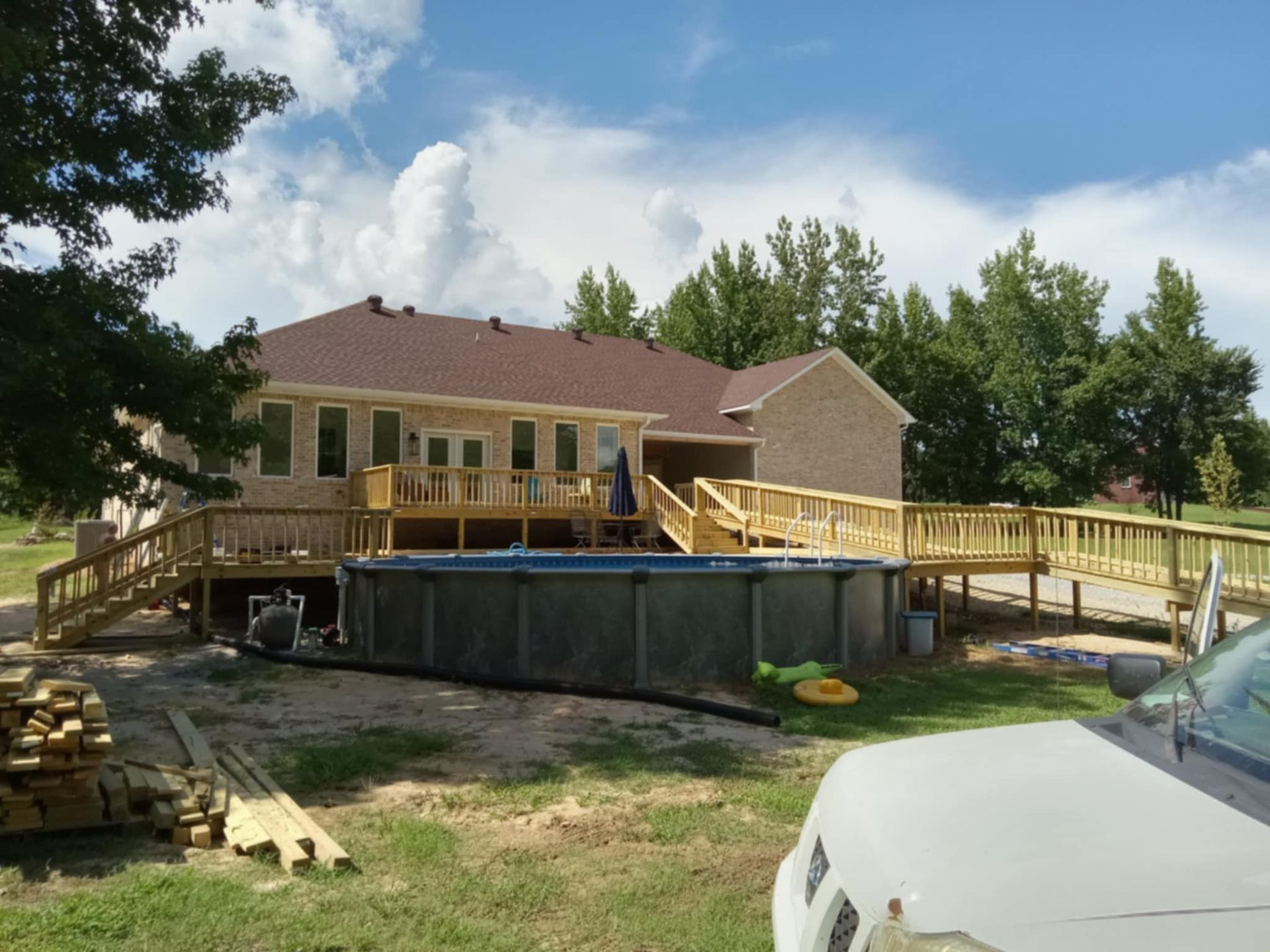House with wooden deck, above-ground pool, and ramp. Sunny day with blue sky and trees.