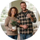 Two people smiling indoors, holding a potted plant and a key, in a bright living room.