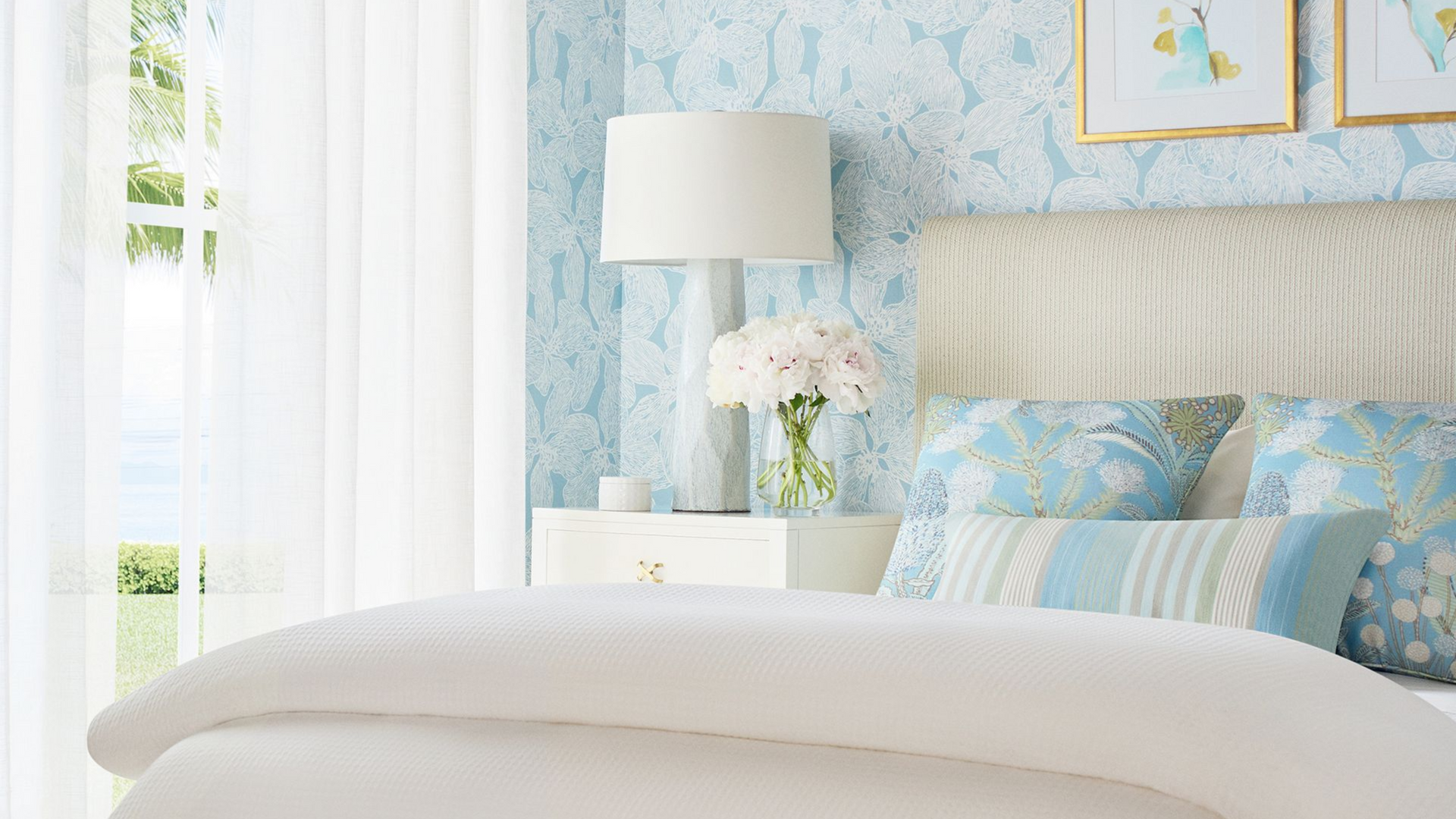 Bedroom with white bedding, blue patterned pillows, and light blue wallpaper; a lamp sits on a white nightstand.