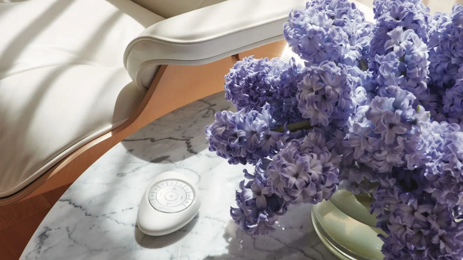 Hunter Douglas Pebble® Remote laying on marble table next to cream chair and vase of lilacs