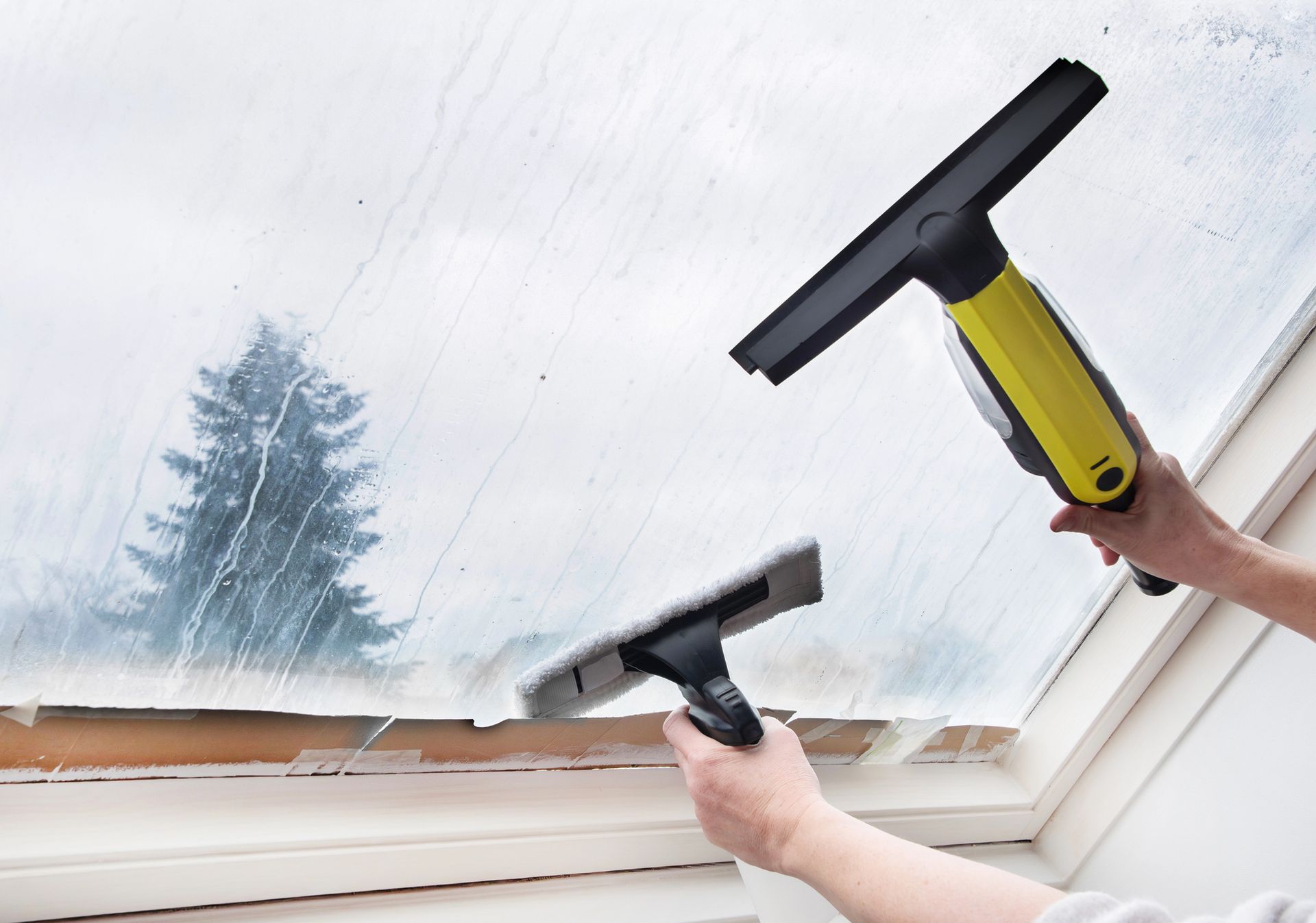 Hands using squeegees to clean a foggy window, with a tree visible outside.