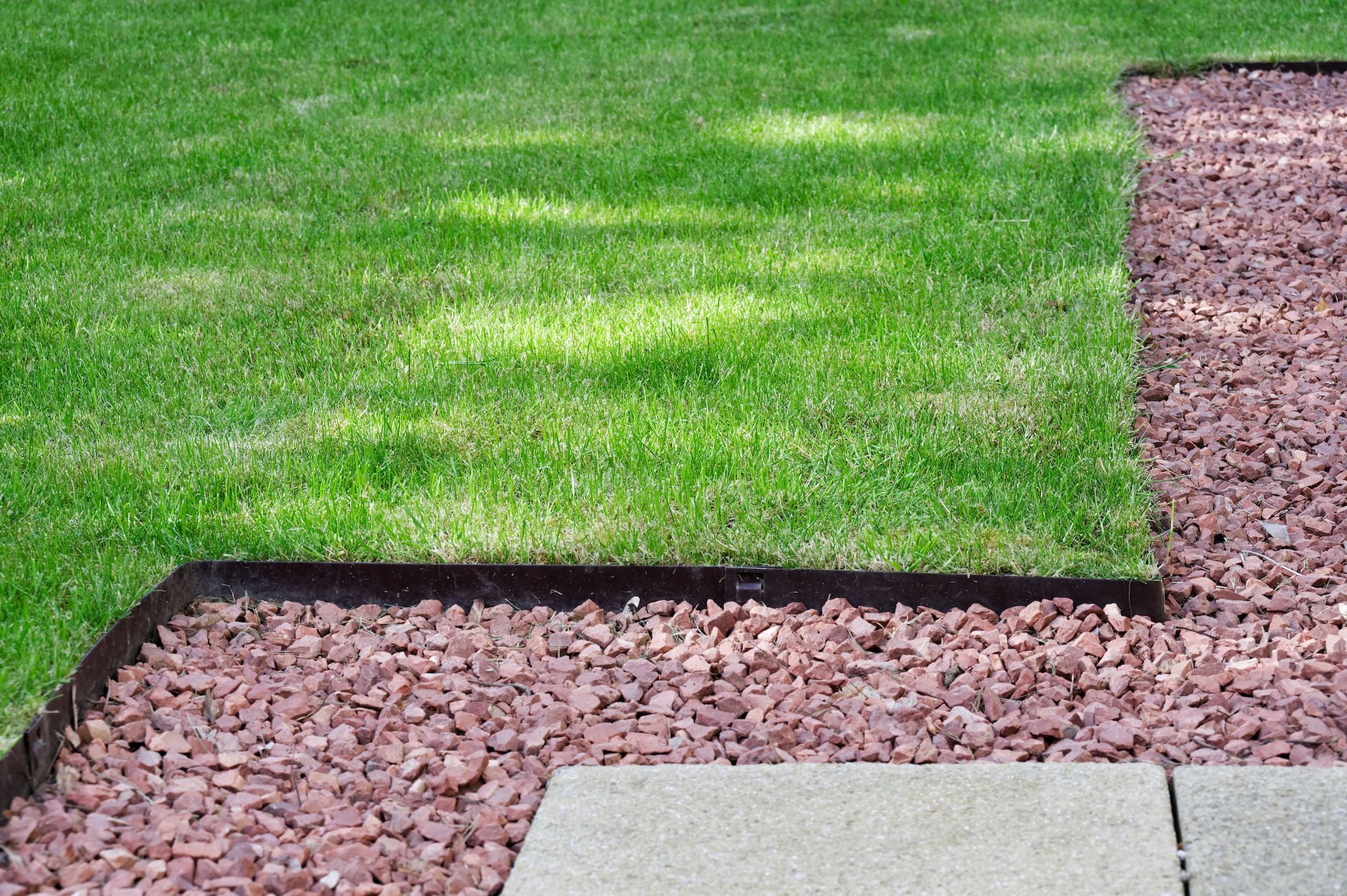 Green lawn bordered by a black edging and pink gravel beside a concrete patio.