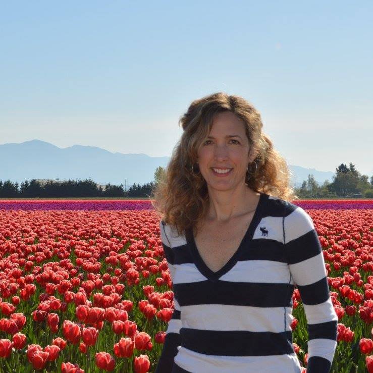 Woman smiling in striped shirt, standing in a field of red tulips with mountains in the background.