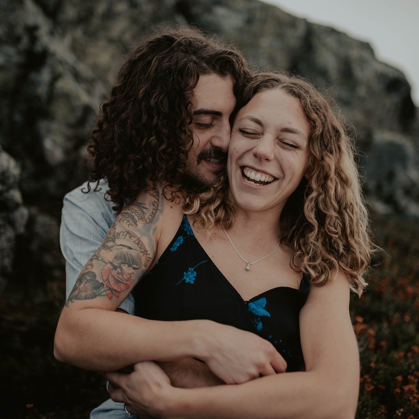 Man hugs laughing woman outdoors; rocky background. He has a tattoo and curly hair. She wears a black dress.