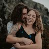 Man hugs laughing woman outdoors; rocky background. He has a tattoo and curly hair. She wears a black dress.