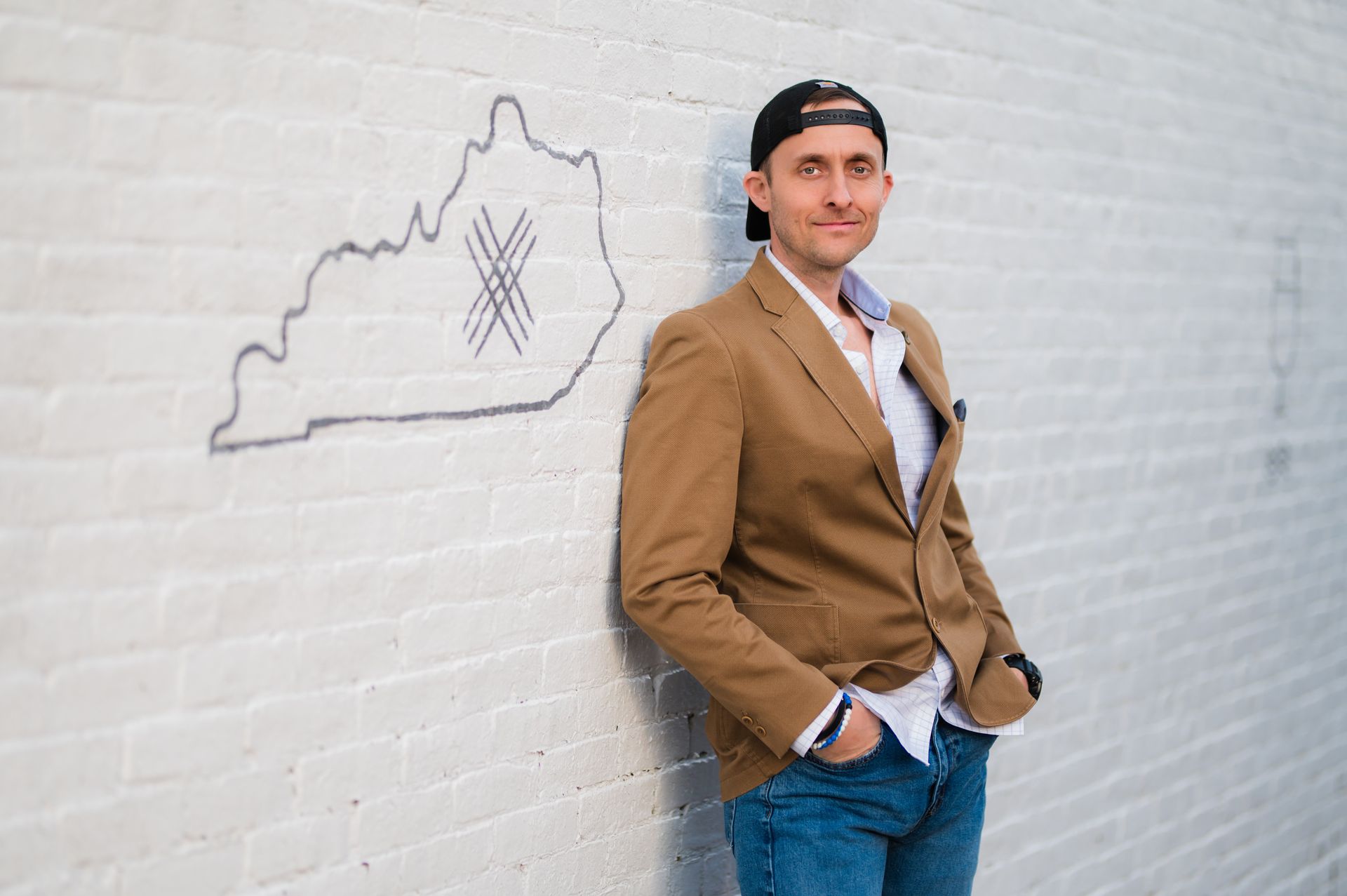 Man in a brown blazer leans against a white brick wall, with a Kentucky outline drawing.