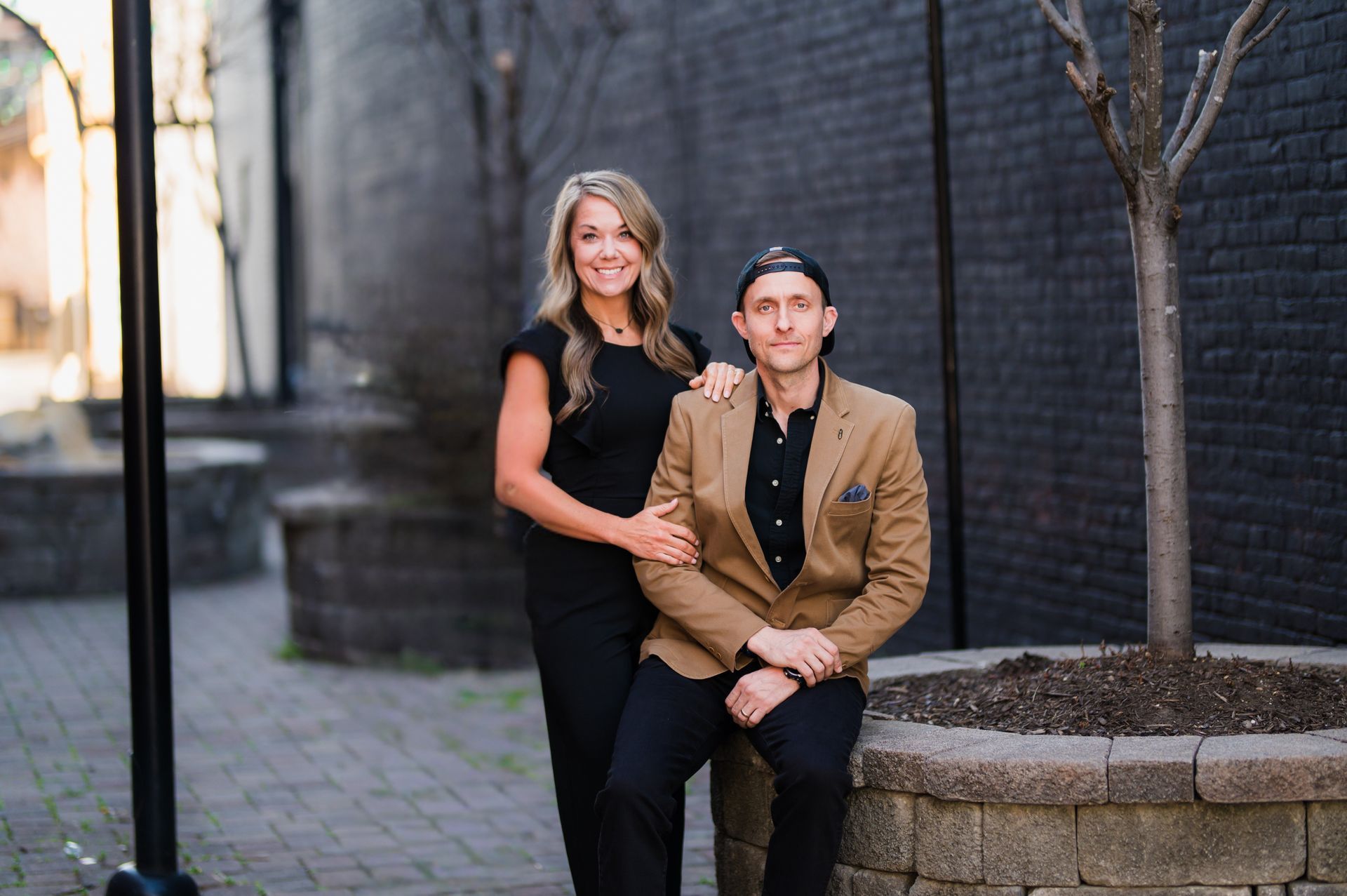 Woman and man pose outdoors in a brick alley. Woman in black dress, man in tan jacket and black cap.