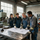 Cuatro trabajadores en un taller revisan planos alrededor de una mesa de metal.