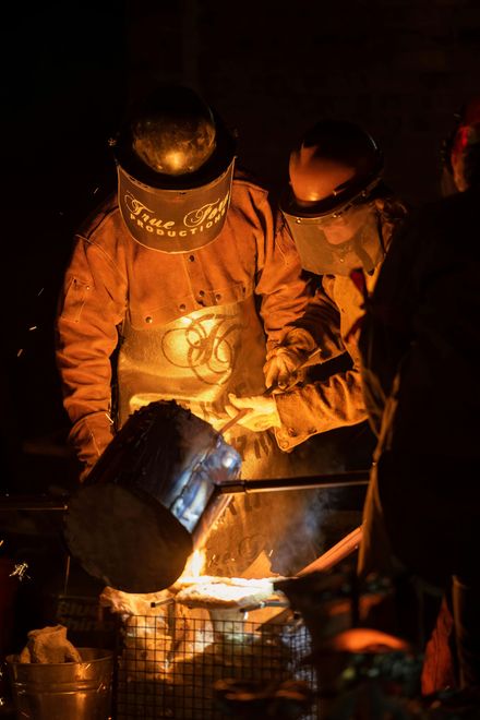 Dos bomberos con trajes naranjas combaten un incendio por la noche, rociando agua sobre las llamas desde un barril de metal.