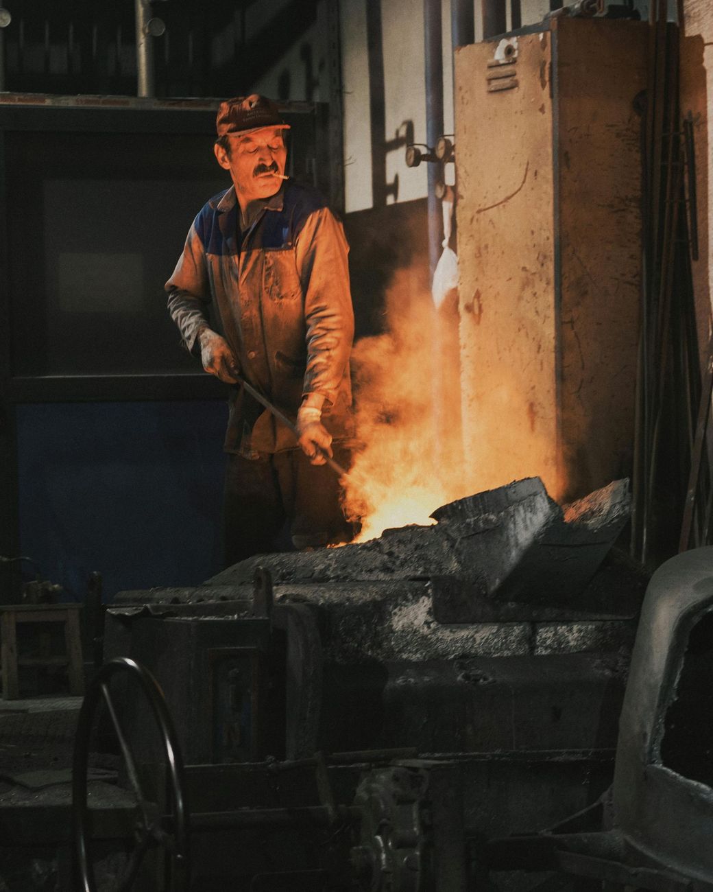 Un obrero de una fundición permanece de pie junto a metal fundido que brilla con un resplandor anaranjado en un oscuro taller industrial.
