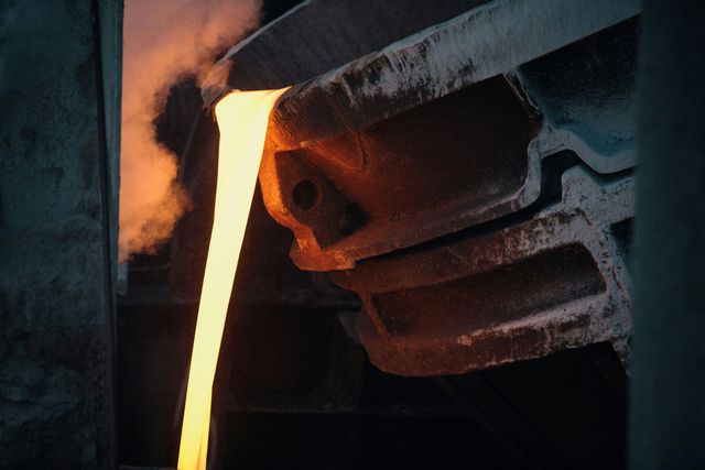Metal fundido vertiéndose desde un horno a un molde, brillando con un tono anaranjado en una fundición oscura.