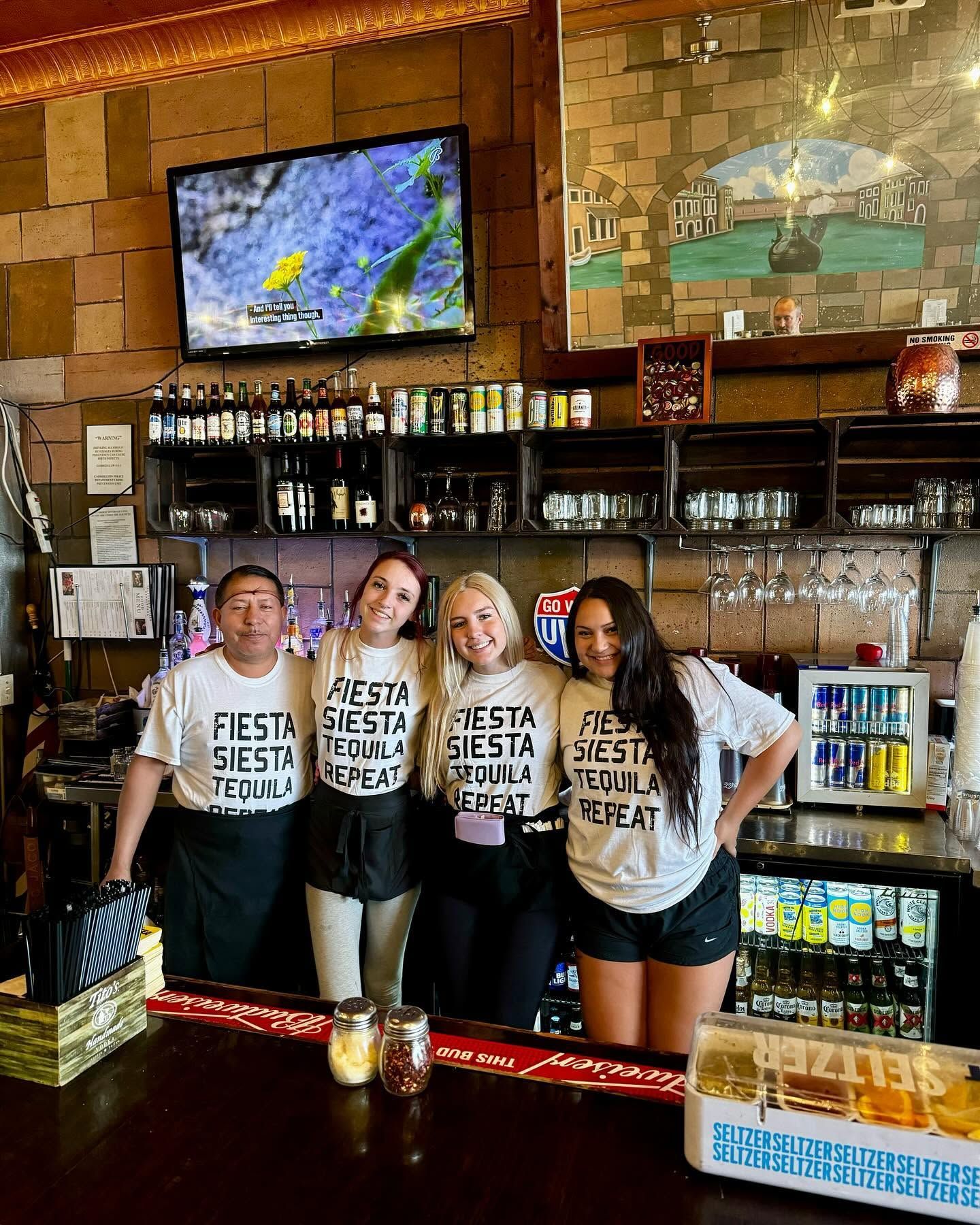 Four people in matching shirts pose behind a bar with liquor bottles and TV screens.