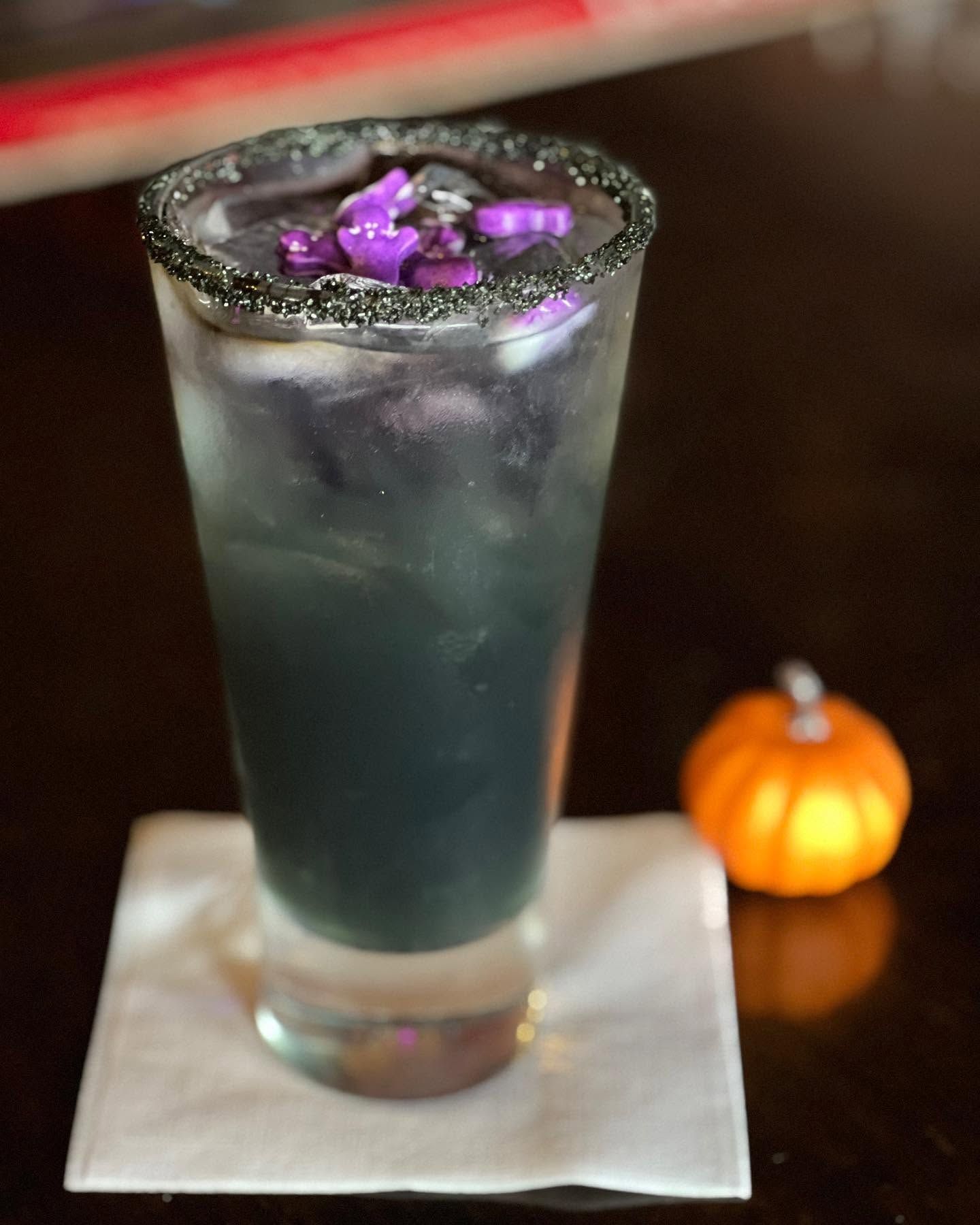 Tall dark cocktail with purple garnish on a napkin beside a small pumpkin on a table