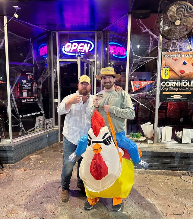 Two people posing in chicken costumes outside a neon-lit shop with an OPEN sign.