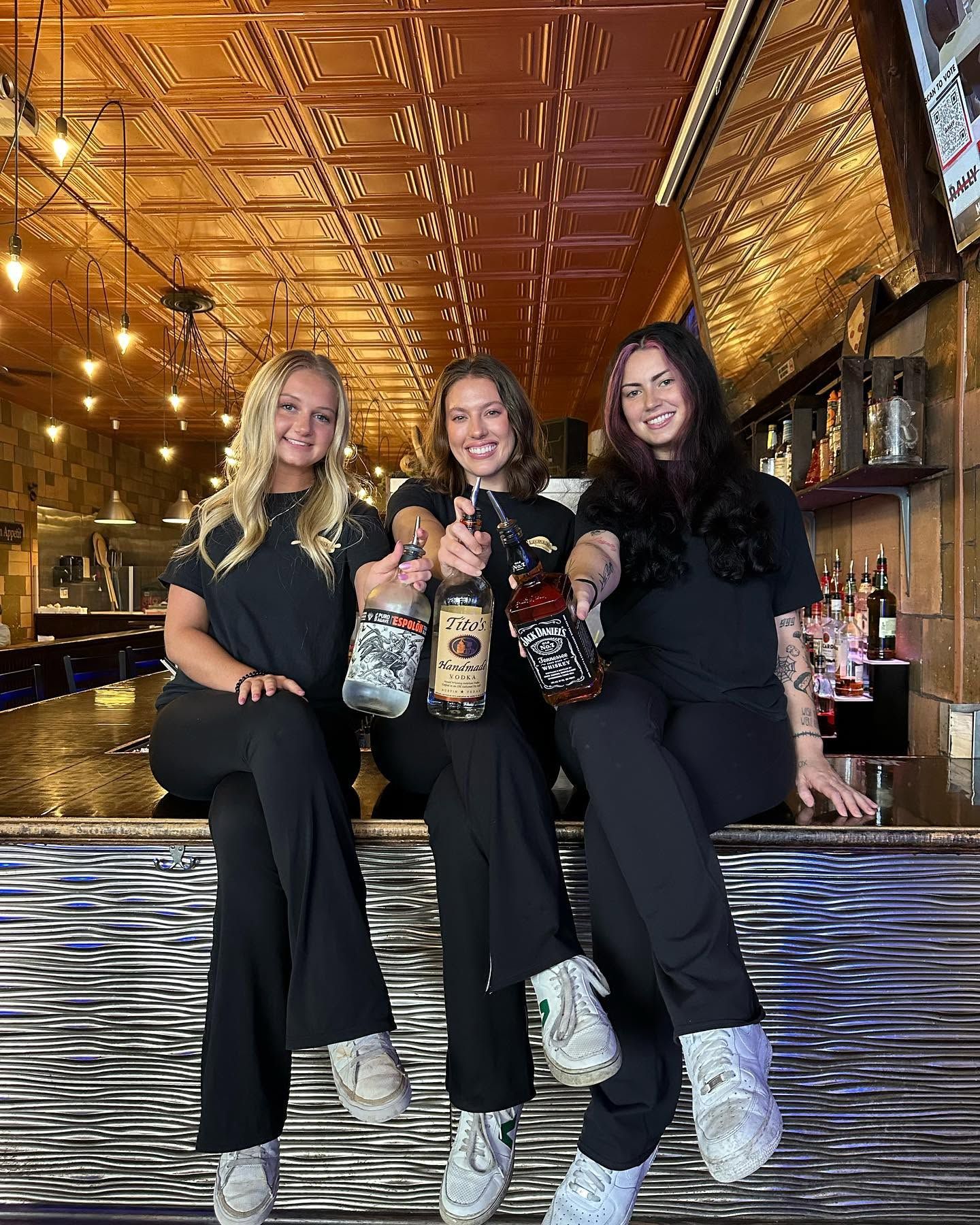 Three friends in black outfits posing with drinks at a warmly lit bar