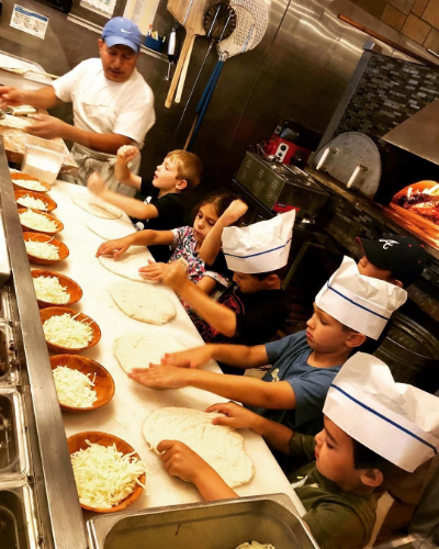 People in chef hats preparing pizzas on a long kitchen counter with sauce, cheese, and toppings