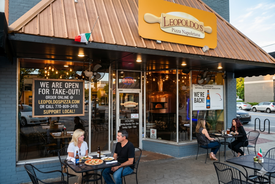 Outdoor café seating at a corner restaurant with people dining and a “We are open for take-out” sign.