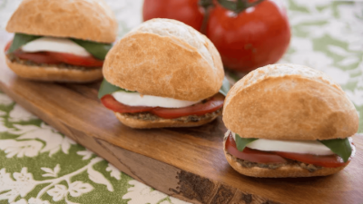 Three mini sandwiches on a wooden board with tomatoes in the background